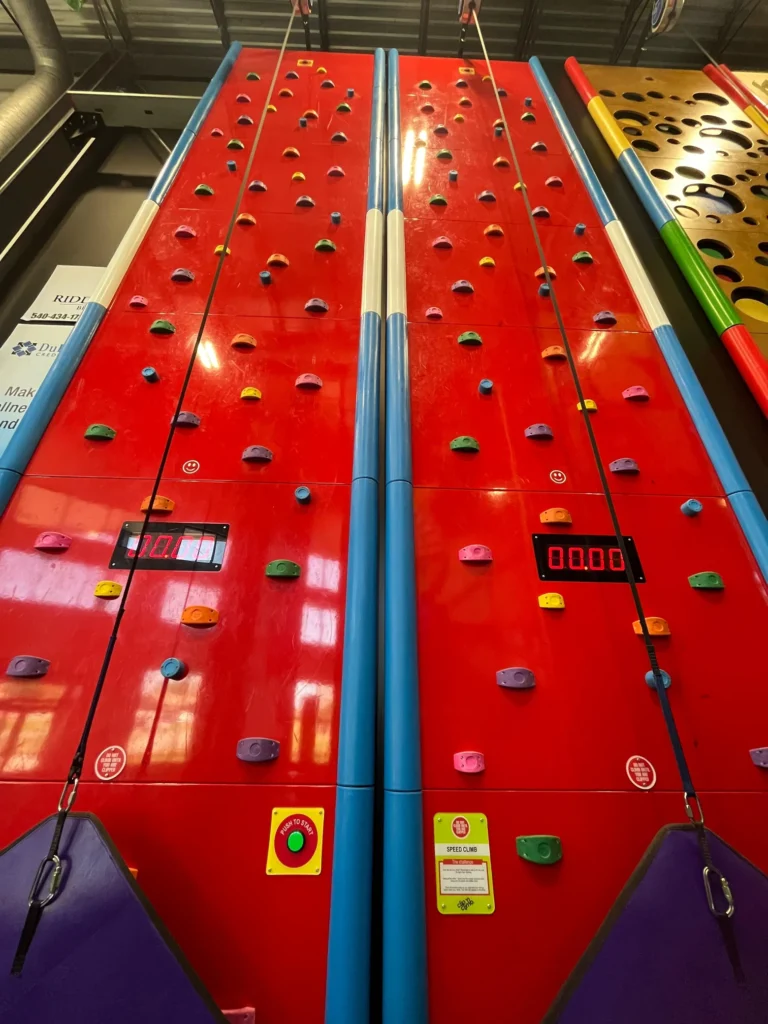 A dual-lane indoor speed climbing wall with colorful holds, digital countdown timers on each lane, and a green start button at the base, designed for head-to-head racing competitions.